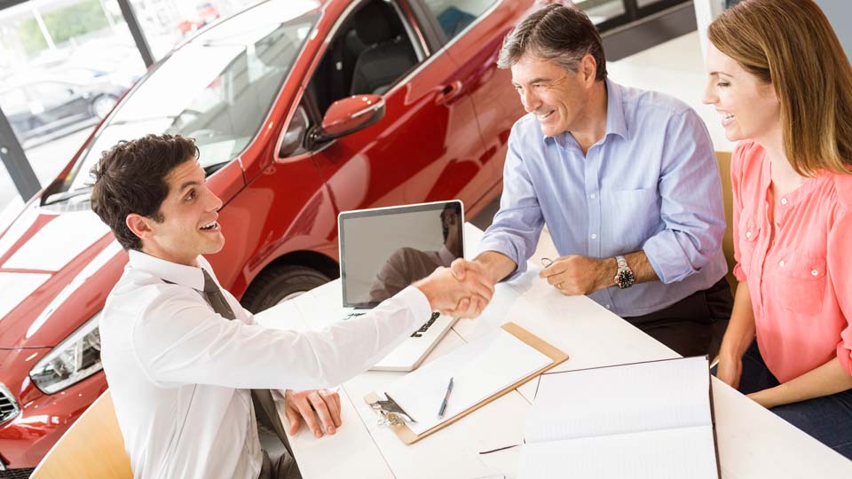 Ein Autoverkäufer schüttelt den Kunden die Hand in einem Autohaus in Aurich, im Hintergrund ist ein rotes Auto zu sehen und auf dem Tisch liegen Unterlagen.